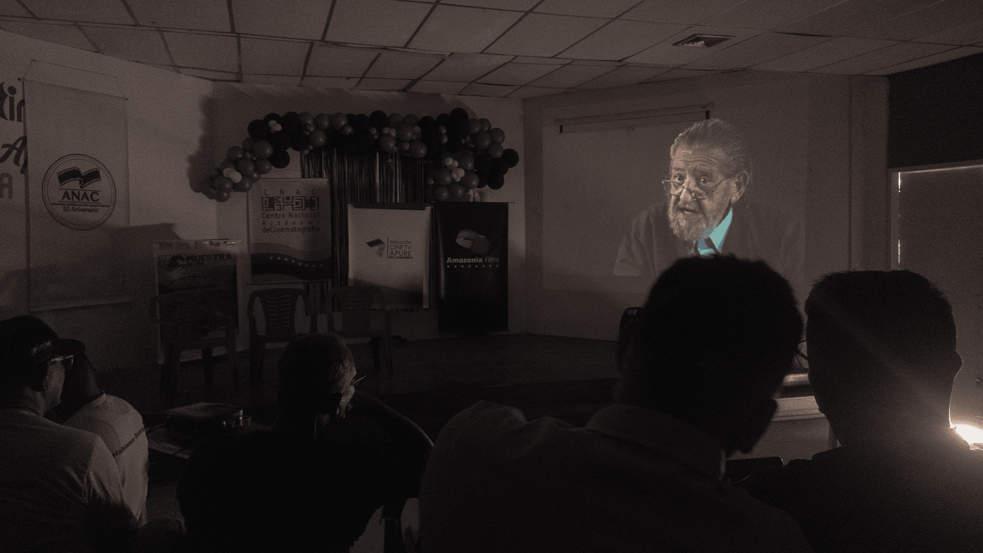 La 3era muestra de cine Apureño y su perspectiva cultural en la actualidad. 5 Audiencia observando una proyección en un evento de Periodismo Constructivo en Apure, con participantes en silueta y una pantalla mostrando a un hombre mayor con barba y gafas.