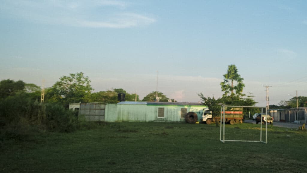 Eliminatorias de la 1era Copa Esmeralda: ¡Se logró! 5 Hierba y árboles en un campo con construcciones modestas y un pequeño arco deportivo en un entorno rural.